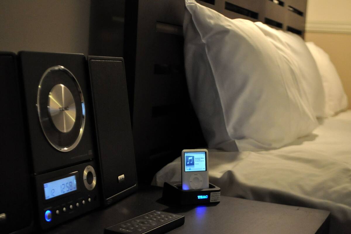 A hotel room with a bed, a bedside table with a clock, a remote control, and an iPod dock.