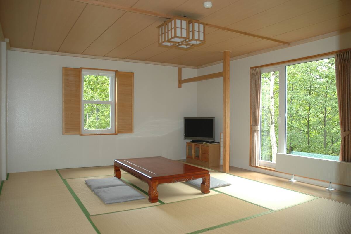 A traditional Japanese room with tatami flooring, a low wooden table, a television, and large windows with curtains.