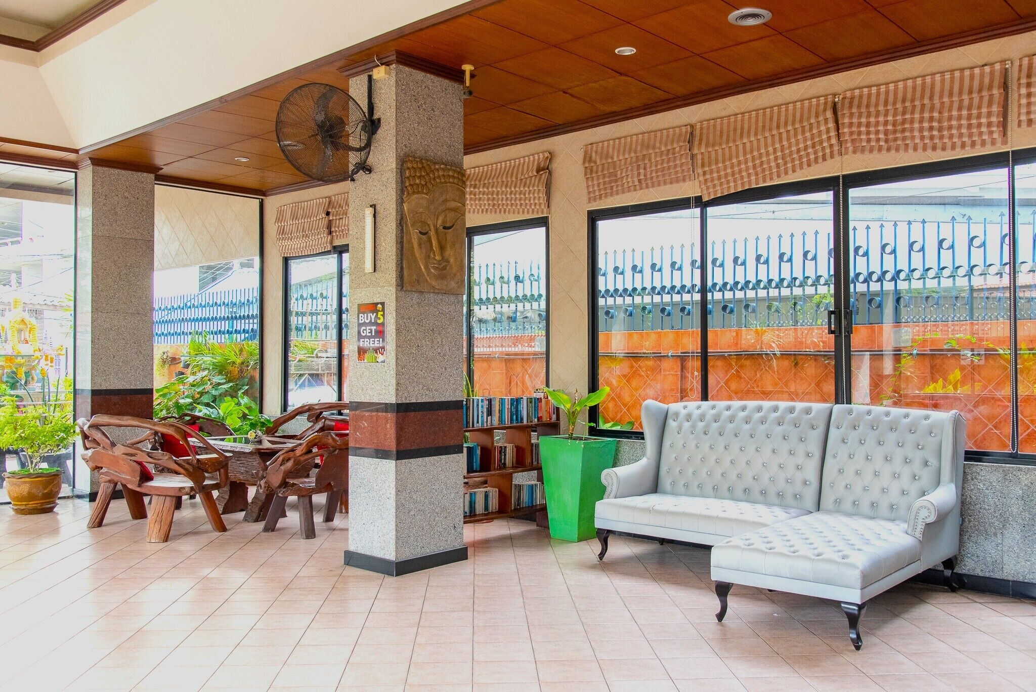 A lobby with a sofa, wooden chairs, a fan, a Buddha statue, a green plant, and a bookshelf.
