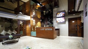 A hotel reception area with a wooden reception desk, a sofa with cushions, and a TV screen displaying a room. - Sleep With Inn (Bangkok)