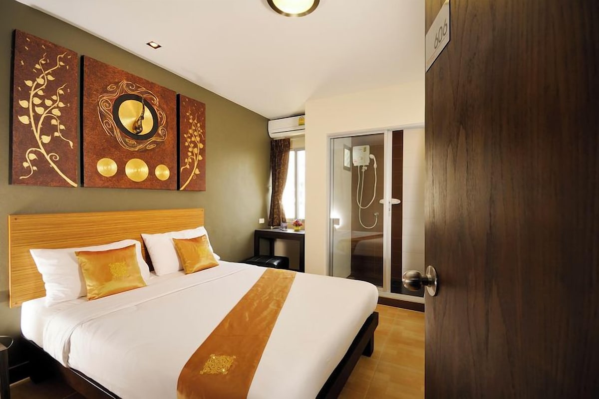A hotel room with a large bed, a wooden headboard, a wall-mounted artwork, a sliding glass door leading to a shower area, and a door with a number sign.