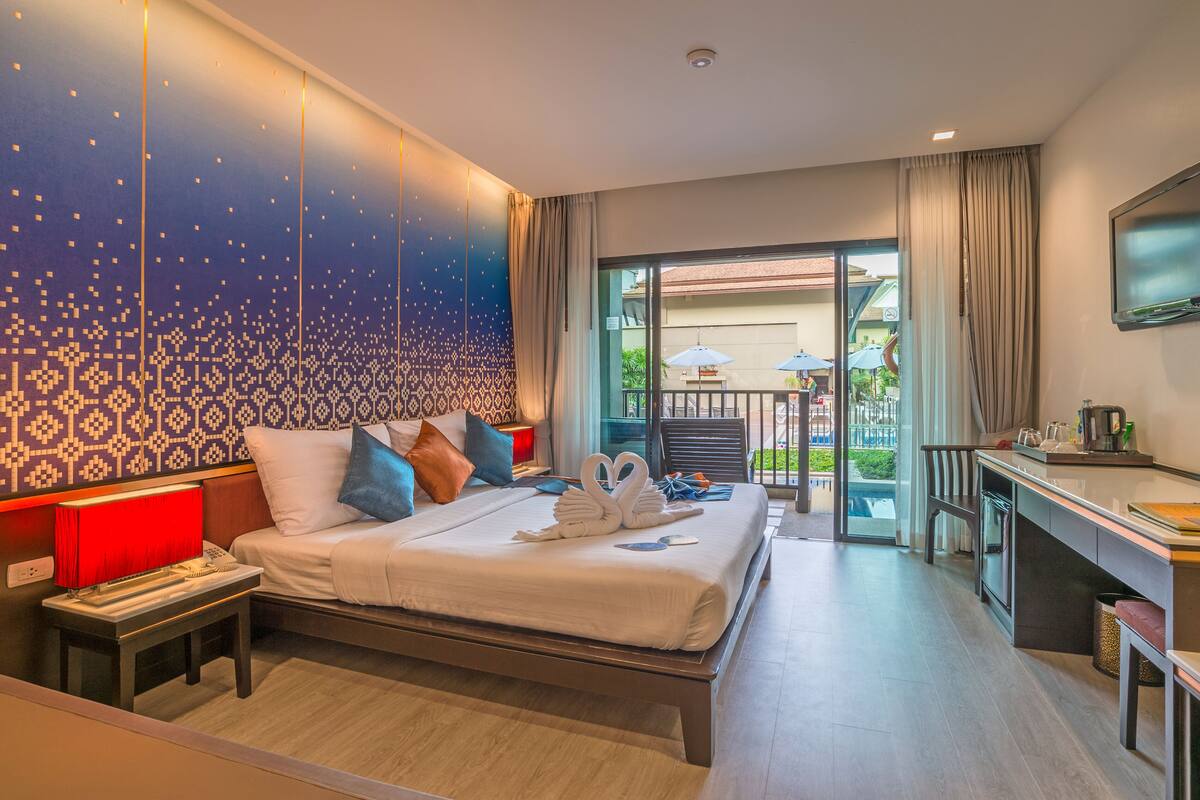 A modern hotel room with a large bed, a desk, and a balcony with outdoor seating.