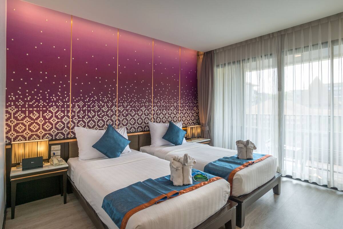A hotel room with two beds, a patterned wall, a nightstand with a lamp, and a view of the city through large windows.