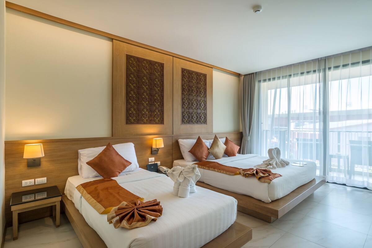 A hotel room with two beds, a nightstand, a lamp, and a view of the outside.