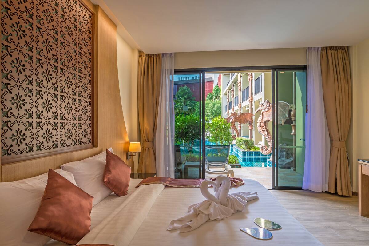 A hotel room with a bed, pillows, a swan-shaped towel, a glass door leading to a balcony with a statue, and a patterned wall.