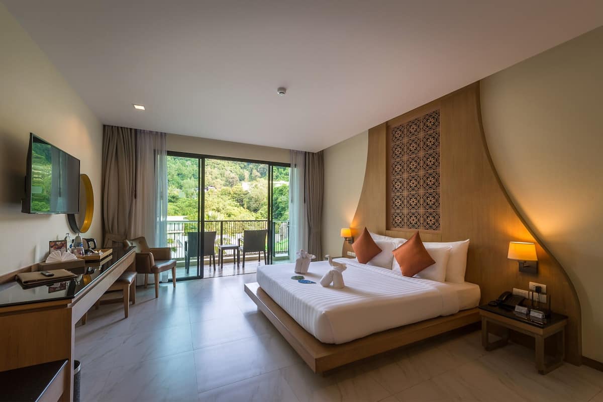 A hotel room with a large bed, a desk, a chair, and a balcony with a view of greenery.