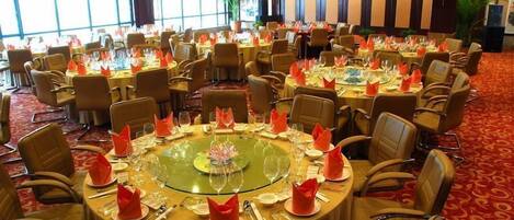 Un salón de banquetes con mesas redondas dispuestas para un evento formal, con centros de mesa de vidrio transparente y servilletas rojas.