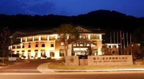 Un hotel de varios pisos con una entrada destacada, un jardín frente al edificio y montañas al fondo.