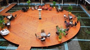 Una terraza circular de madera con mesas y sillas, rodeada de plantas en macetas y un pilar central.