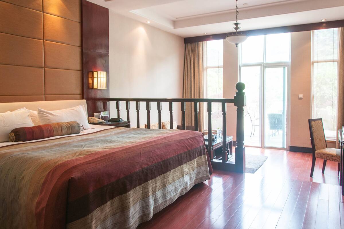 A hotel room with a large bed, a wooden headboard, a balcony with a chair, and a view of the outdoors.