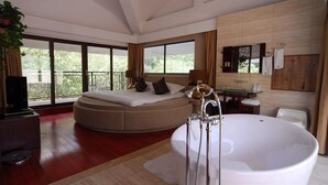 Een moderne hotelkamer met een groot bed, een zithoek, een badkuip en een balkon met uitzicht op groen.