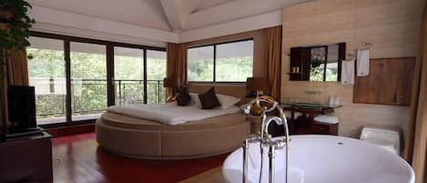 A modern hotel room with a large bed, a separate seating area, a bathtub, and a balcony with a view of greenery.
