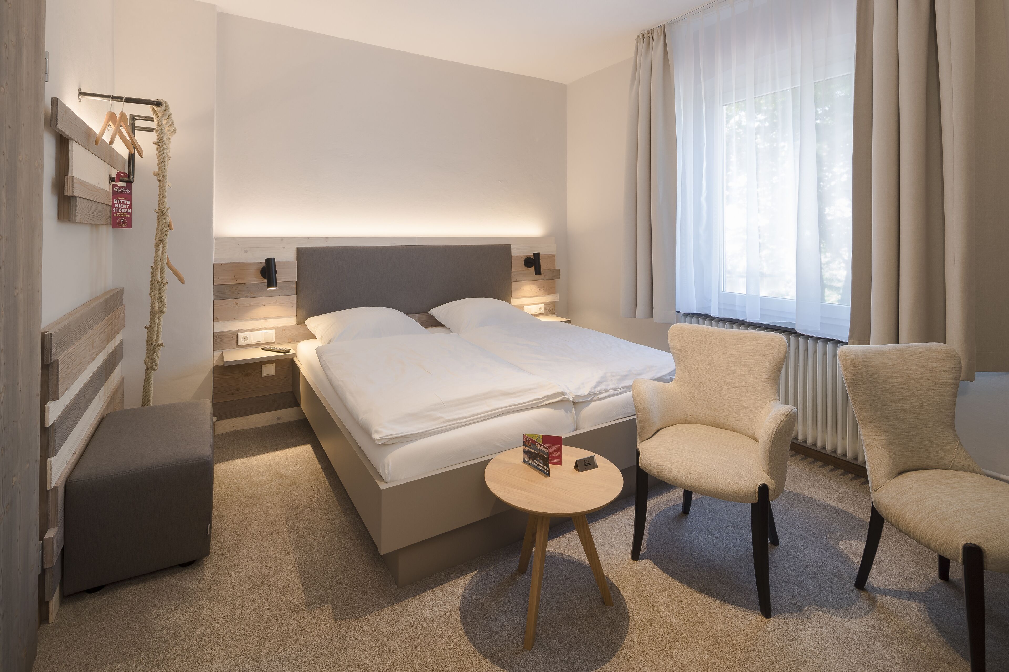 A modern hotel room with a large bed, bedside tables, a small round table, a chair, and a window with curtains.
