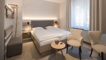 A modern hotel room with a large bed, bedside tables, a small round table, a chair, and a window with curtains.