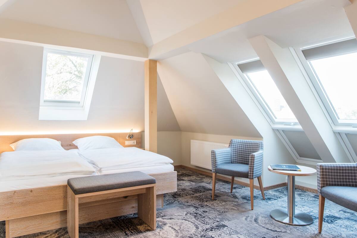 A bedroom with a bed, bedside tables, a bench, a small round table, and two chairs. There are skylights and a window providing natural light.