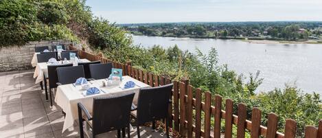 An outdoor dining area with tables and chairs, overlooking a body of water and a distant town.