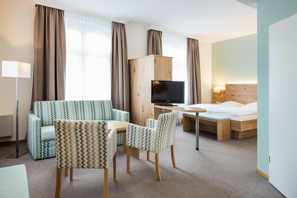 A hotel room with a sofa, two chairs, a bed, a TV, and a wooden cabinet.
