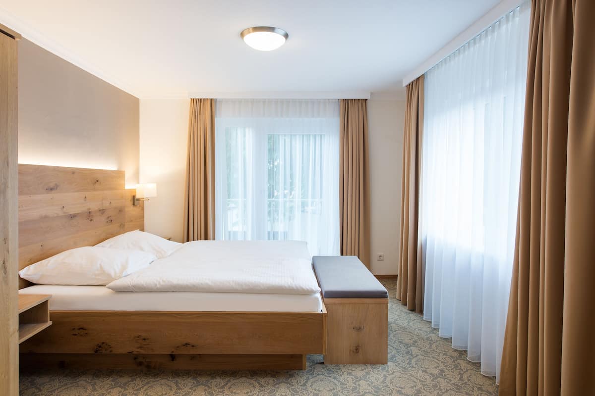 A hotel room with a wooden bed, a bench, a bedside lamp, and sheer curtains.