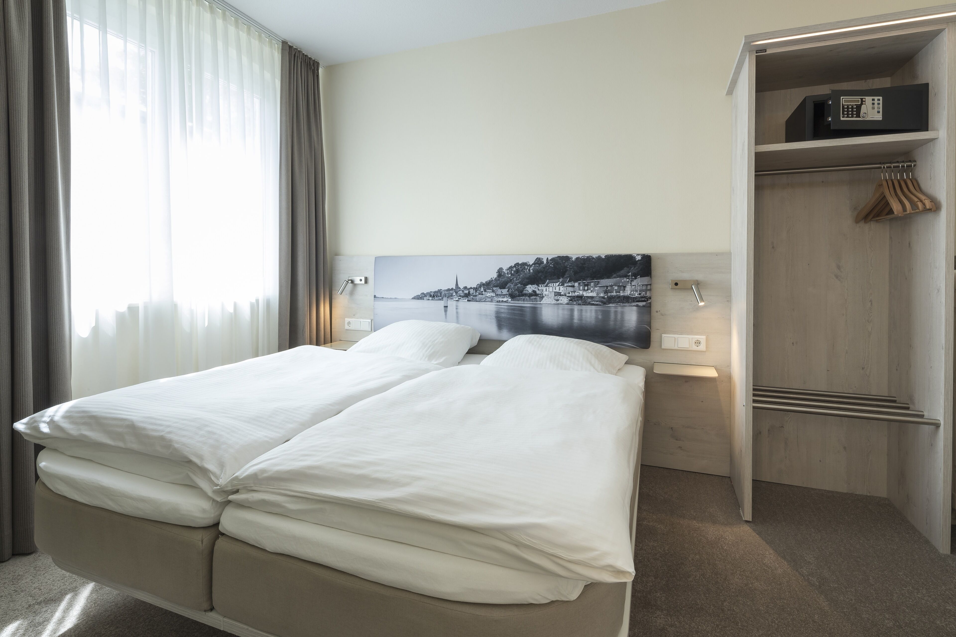 A hotel room with a bed, a wooden wardrobe, a painting of a coastal scene, and a window with curtains.