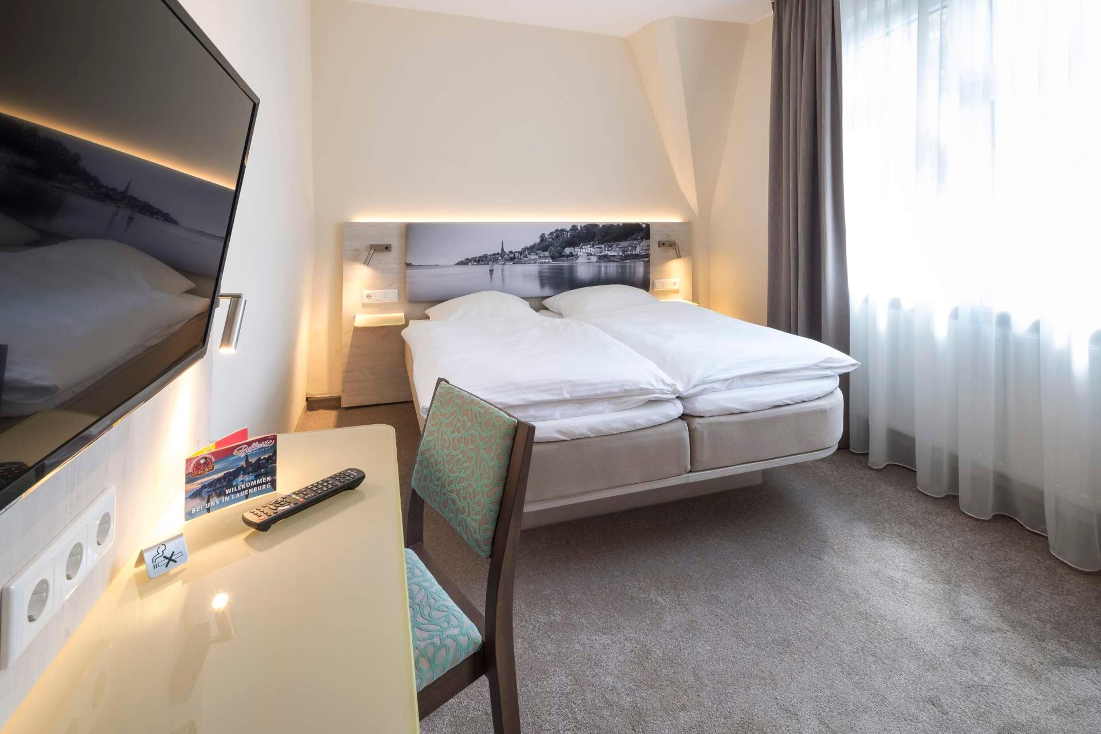 A hotel room with a bed, a desk with a chair, a TV, and a painting on the wall.