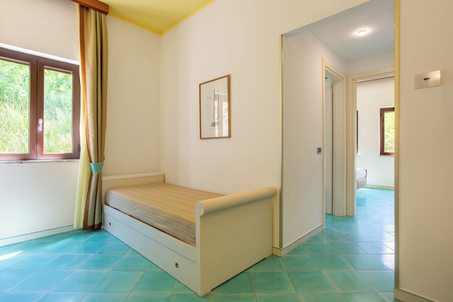 A small room with a bed, a window with curtains, a framed picture on the wall, and a door leading to another room.