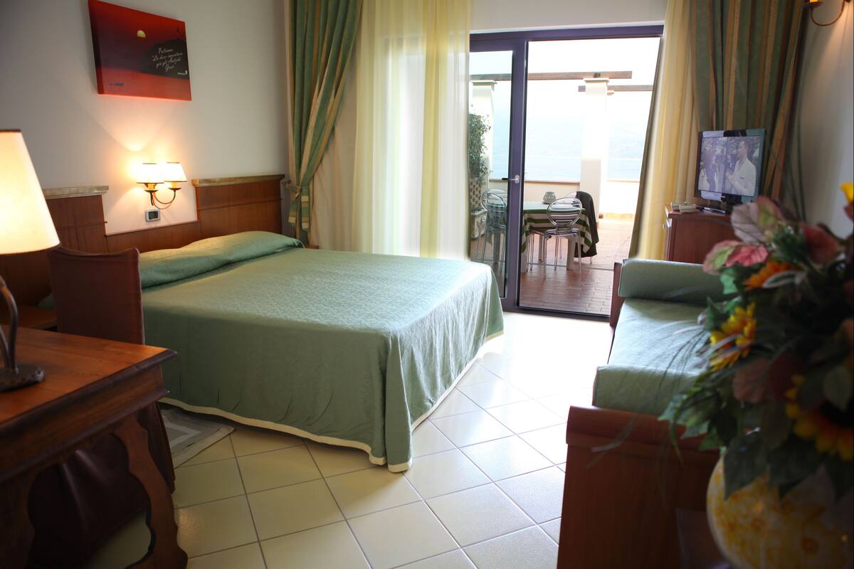A hotel room with a bed, a desk with a lamp, a TV, and a balcony with a table and chairs.