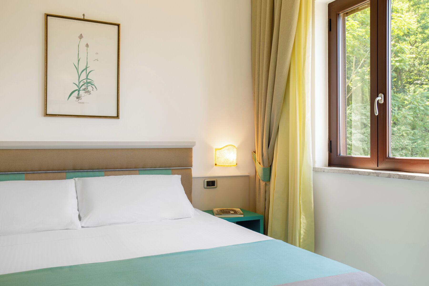 A neatly made bed with white and teal bedding, a framed botanical illustration on the wall, a bedside table with a lamp, and a window with curtains.