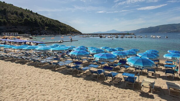 A beach with rows of sun loungers and umbrellas, a pier extending into the water, and mountains in the background.