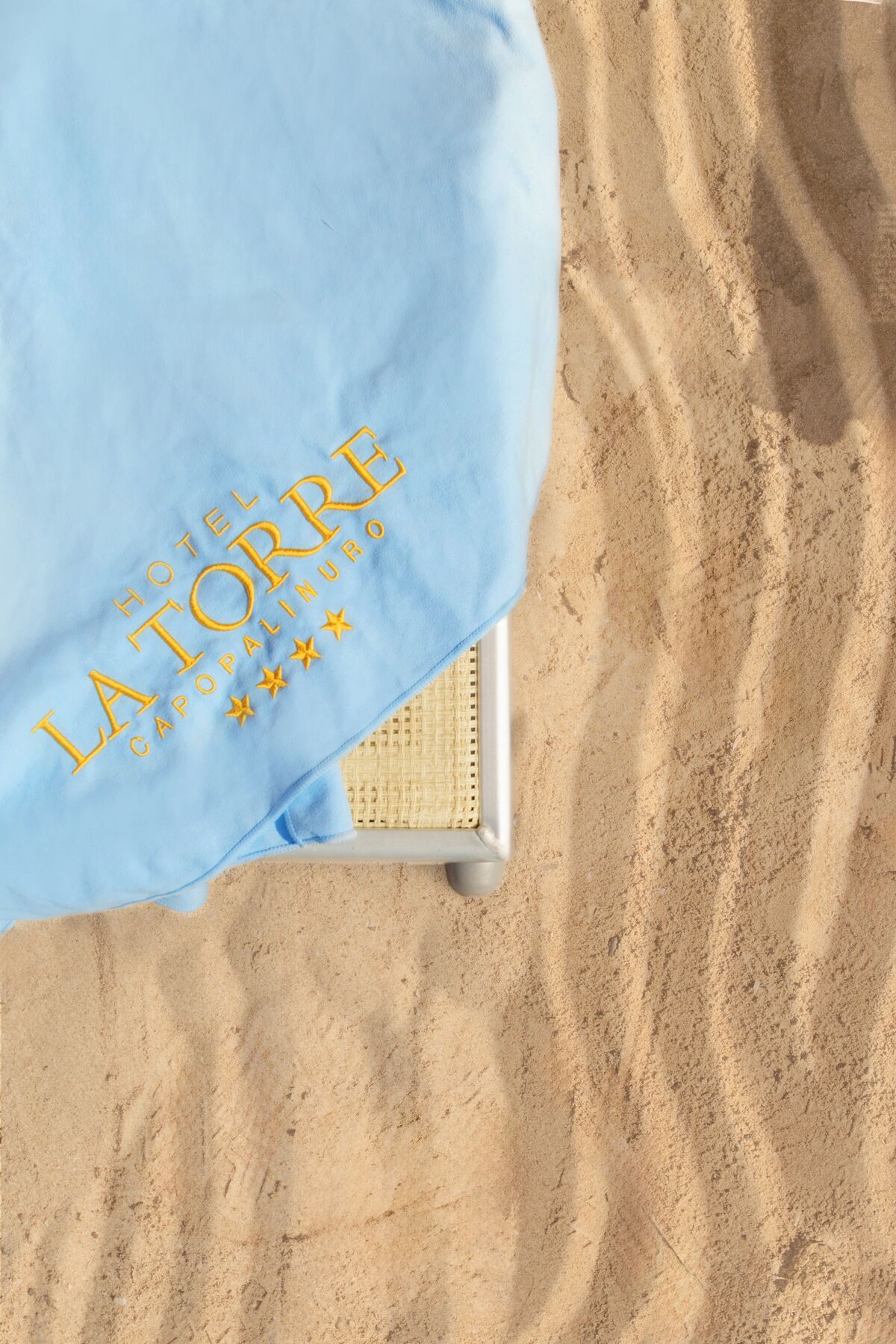 A blue pillow with "HOTEL LATORRE CAPOPALIO" and three stars embroidered on it, placed on a white chair on a sandy surface.