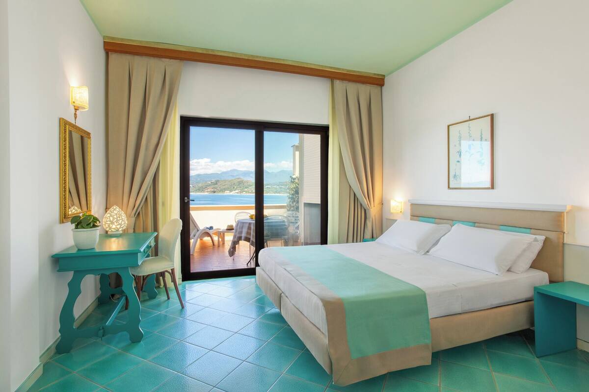 A hotel room with a bed, a desk, a chair, a table, and a view of the sea.