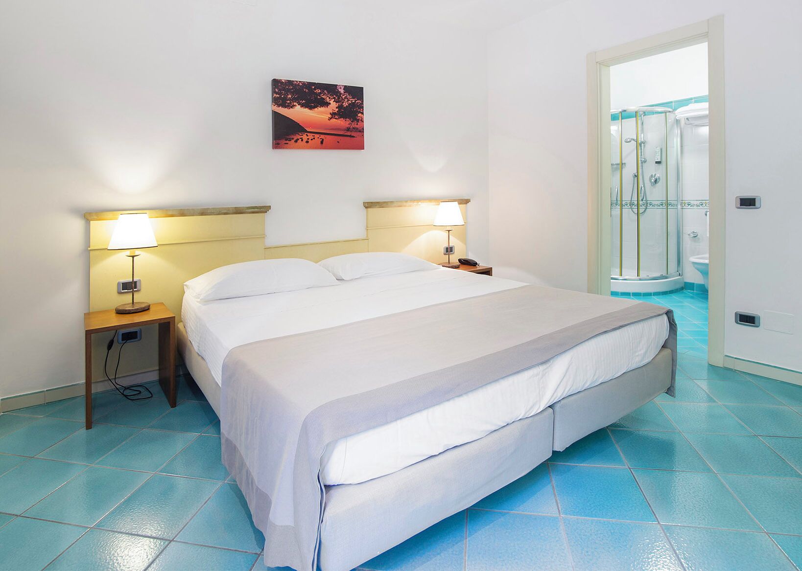 Una camera d'albergo con un letto, due lampade sui comodini, un quadro appeso al muro e un angolo doccia visibile attraverso una porta aperta.