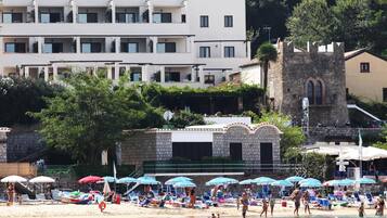 A beach with sun loungers and umbrellas, a stone building, and a tower.
