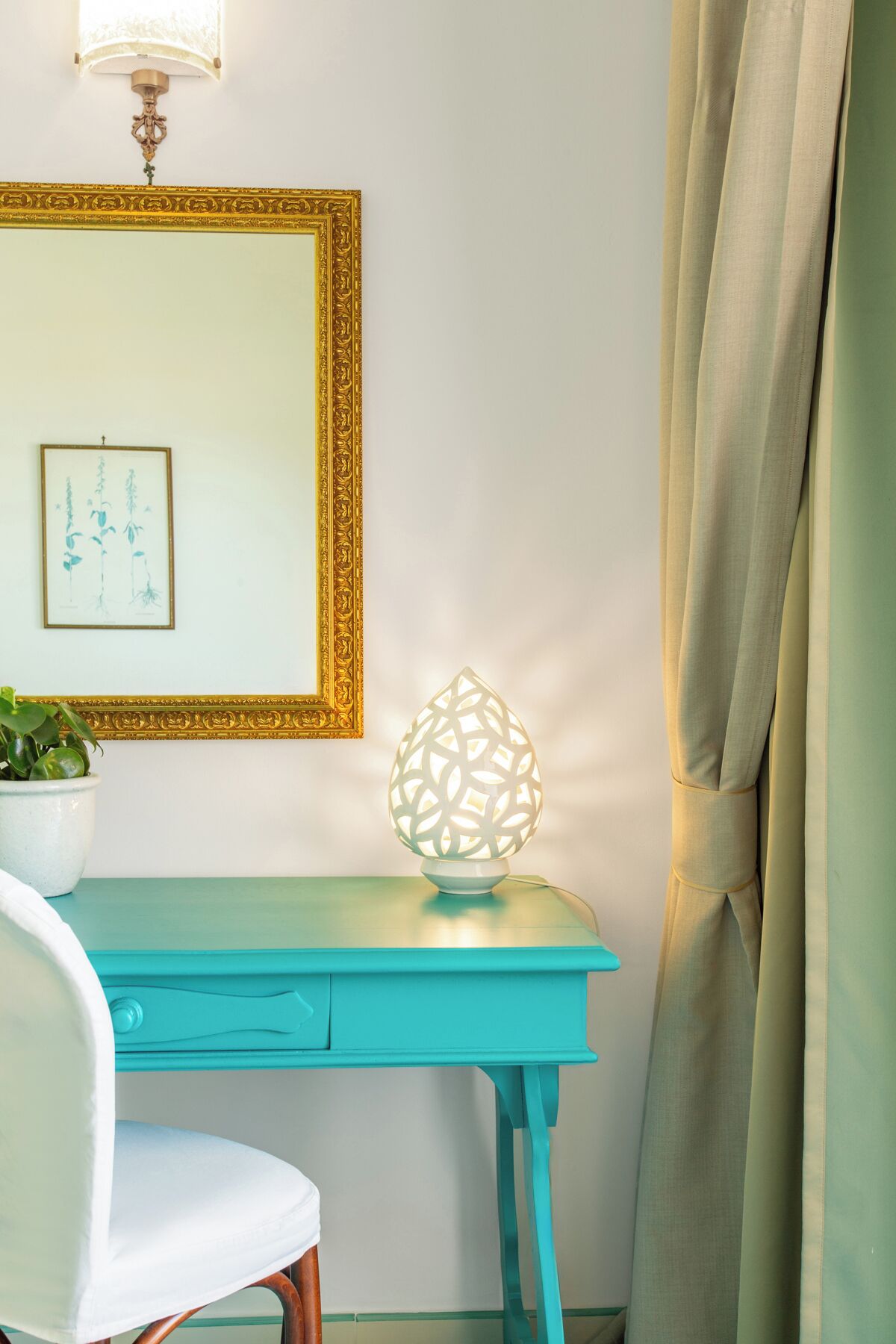 Un escritorio color turquesa con una silla blanca, una lámpara decorativa, una obra de arte enmarcada y una planta en maceta.