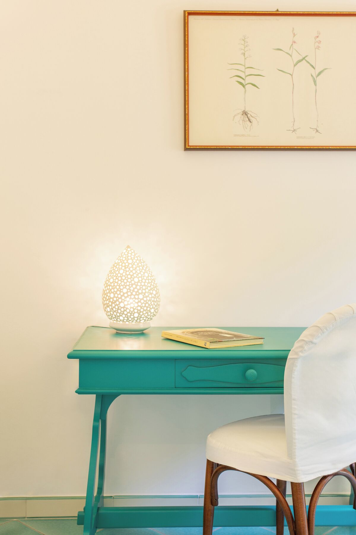Et teal-farvet skrivebord med en hvid stol, en dekorativ lampe og et indrammet botanisk tryk på væggen.