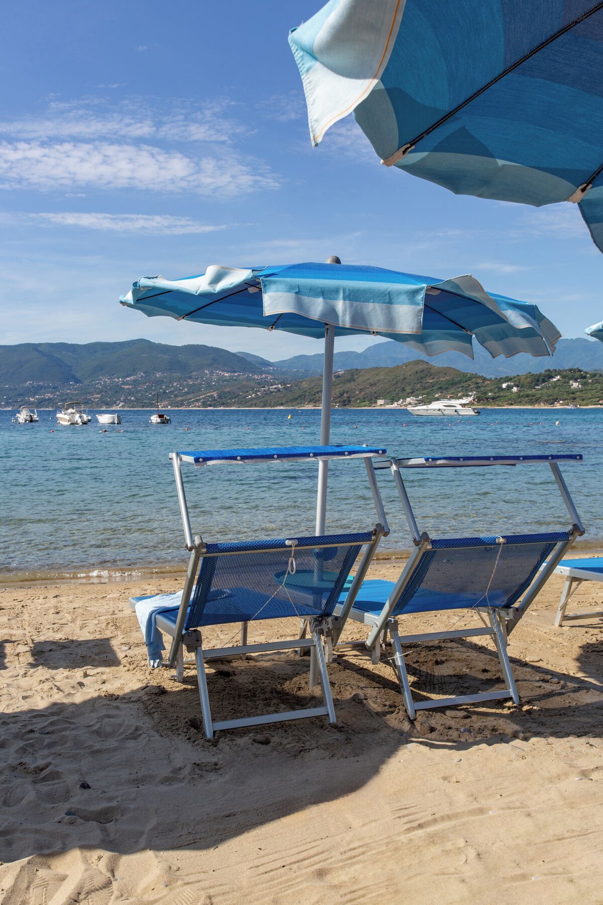 Ein Strand mit zwei blauen Liegestühlen und einem großen blau-weißen Sonnenschirm. Im Wasser sind Boote zu sehen, und im Hintergrund eine gebirgige Landschaft.