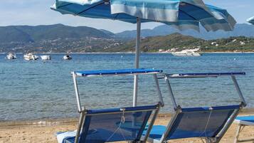 Ein Strand mit zwei blauen Liegestühlen und einem großen blau-weißen Sonnenschirm. Im Wasser sind Boote zu sehen, und im Hintergrund eine gebirgige Landschaft.