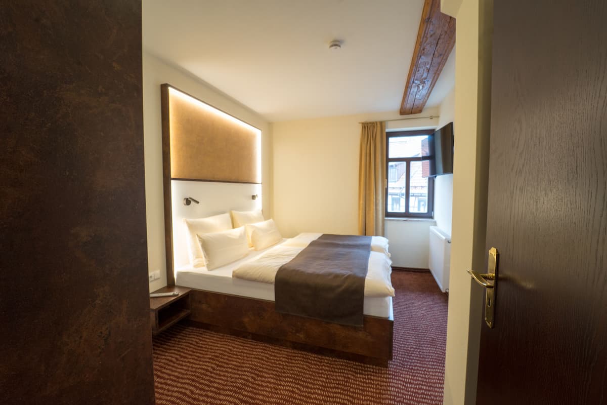 Habitación de hotel con una cama grande, cabecera de madera, ventana con cortinas y una puerta de madera.