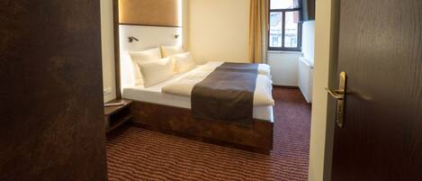 Habitación de hotel con una cama grande, cabecera de madera, ventana con cortinas y una puerta de madera.
