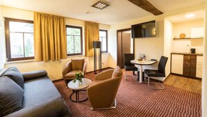 A hotel room with a sofa, two armchairs, a small round table, a flat-screen TV, a kitchen area with a microwave, and a large window with curtains. - Gasthaus Goldener Hirsch (Suhl)