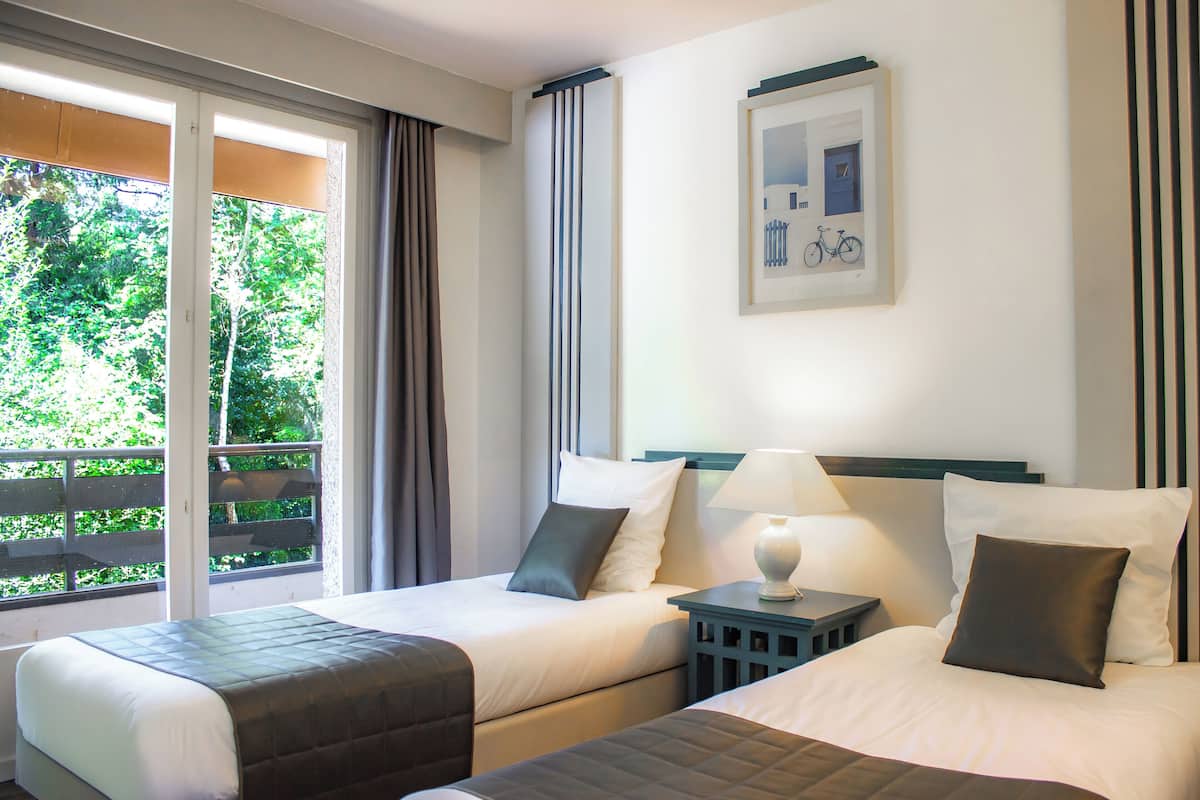A hotel room with two beds, a window with curtains, a framed picture on the wall, and a nightstand with a lamp.