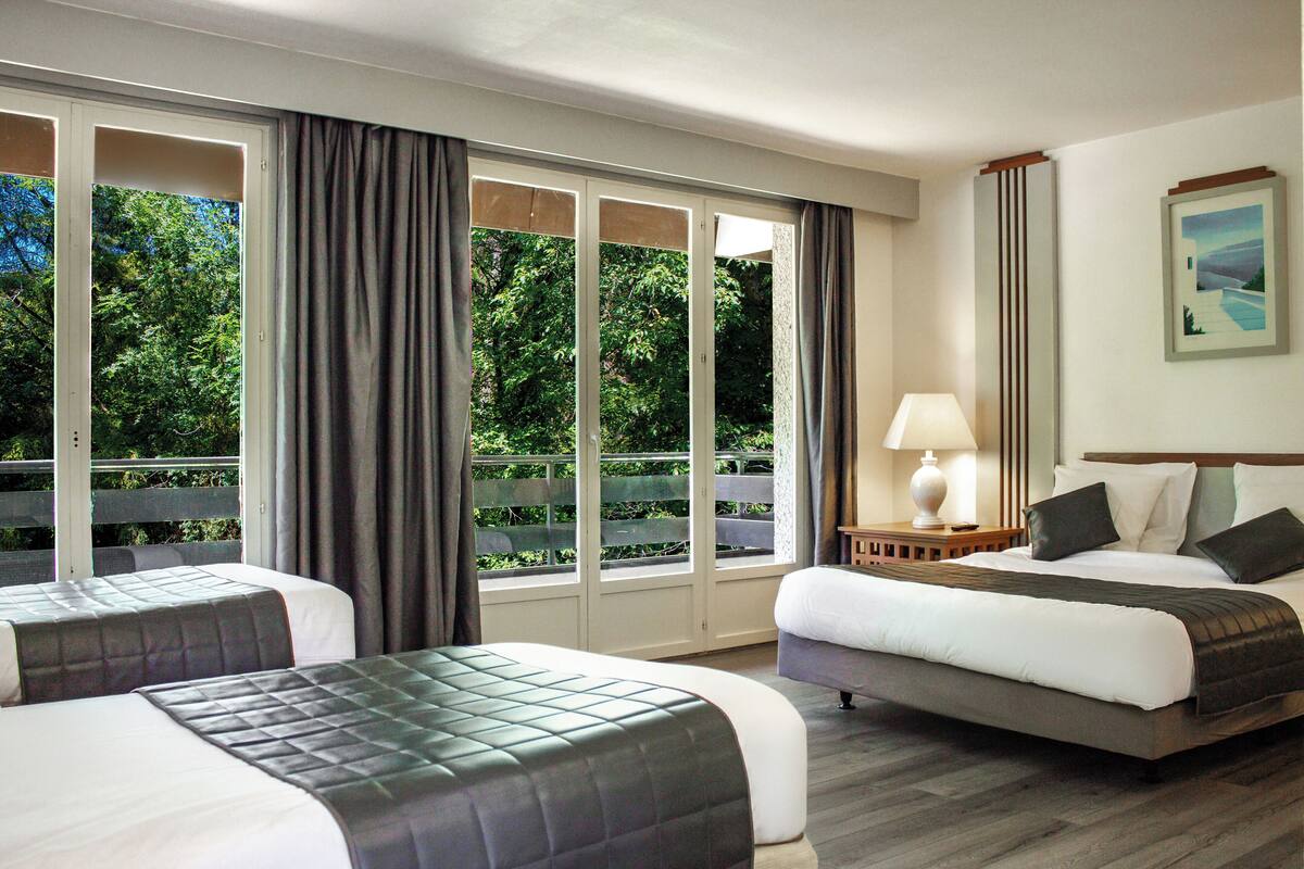 A hotel room with two beds, a large window with curtains, a nightstand with a lamp, and a framed picture on the wall.