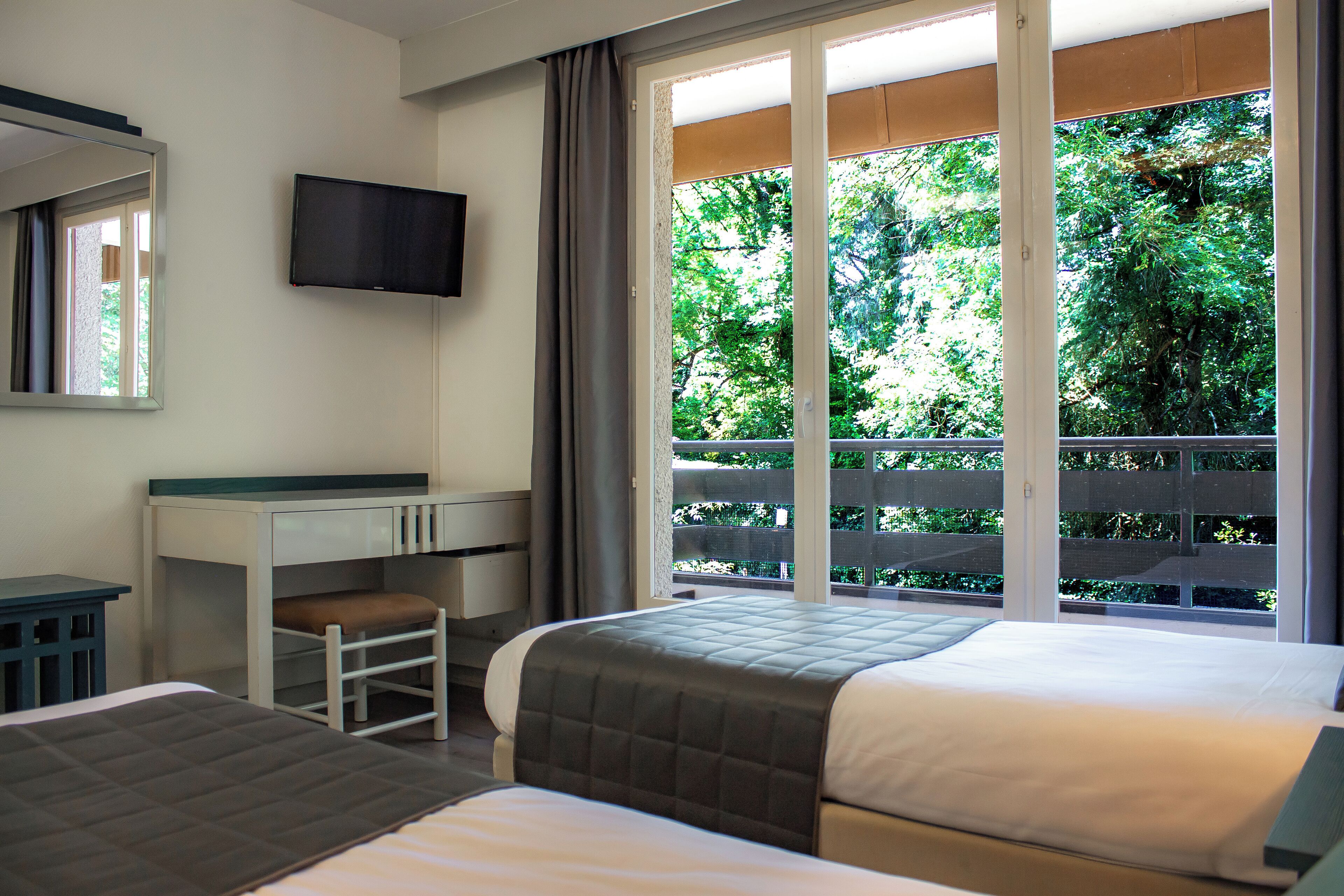 A hotel room with two beds, a desk, a chair, a television, and a balcony with a view of greenery.