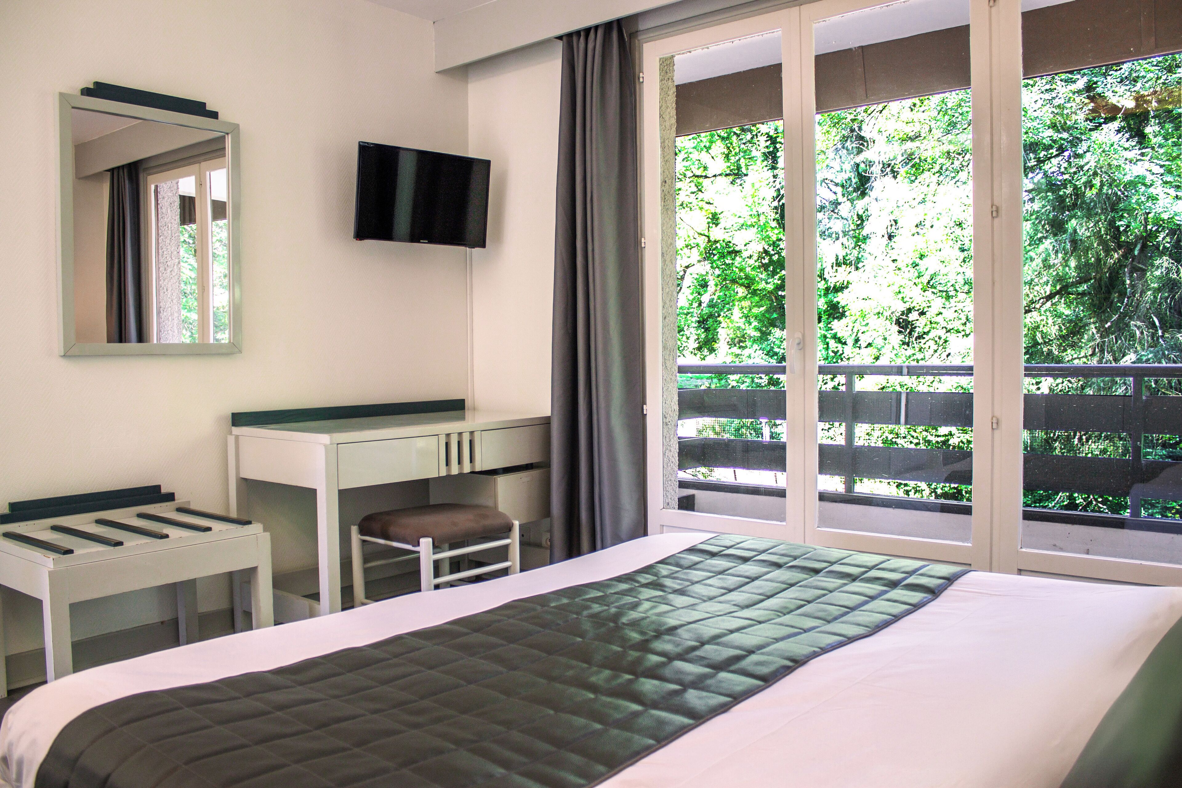 Hotelkamer met een bed, bureau, stoel, televisie en balkon met uitzicht op bomen.