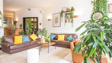 Een moderne woonkamer met een bruine bank, gele en blauwe kussens, een bijzettafeltje en verschillende planten.