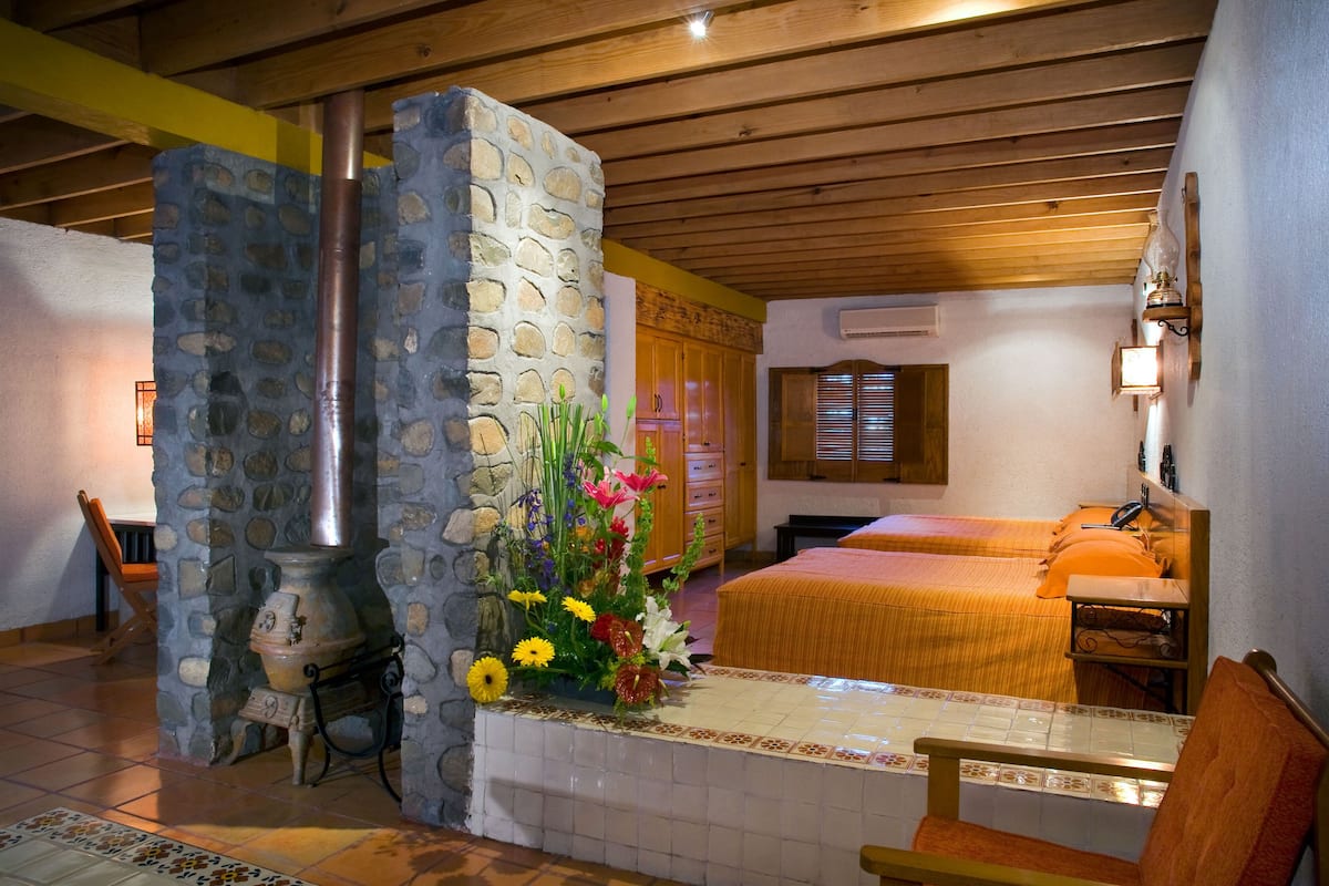 Una habitación acogedora con chimenea de piedra, una cama con ropa de cama naranja, techo de madera y suelo de baldosas.