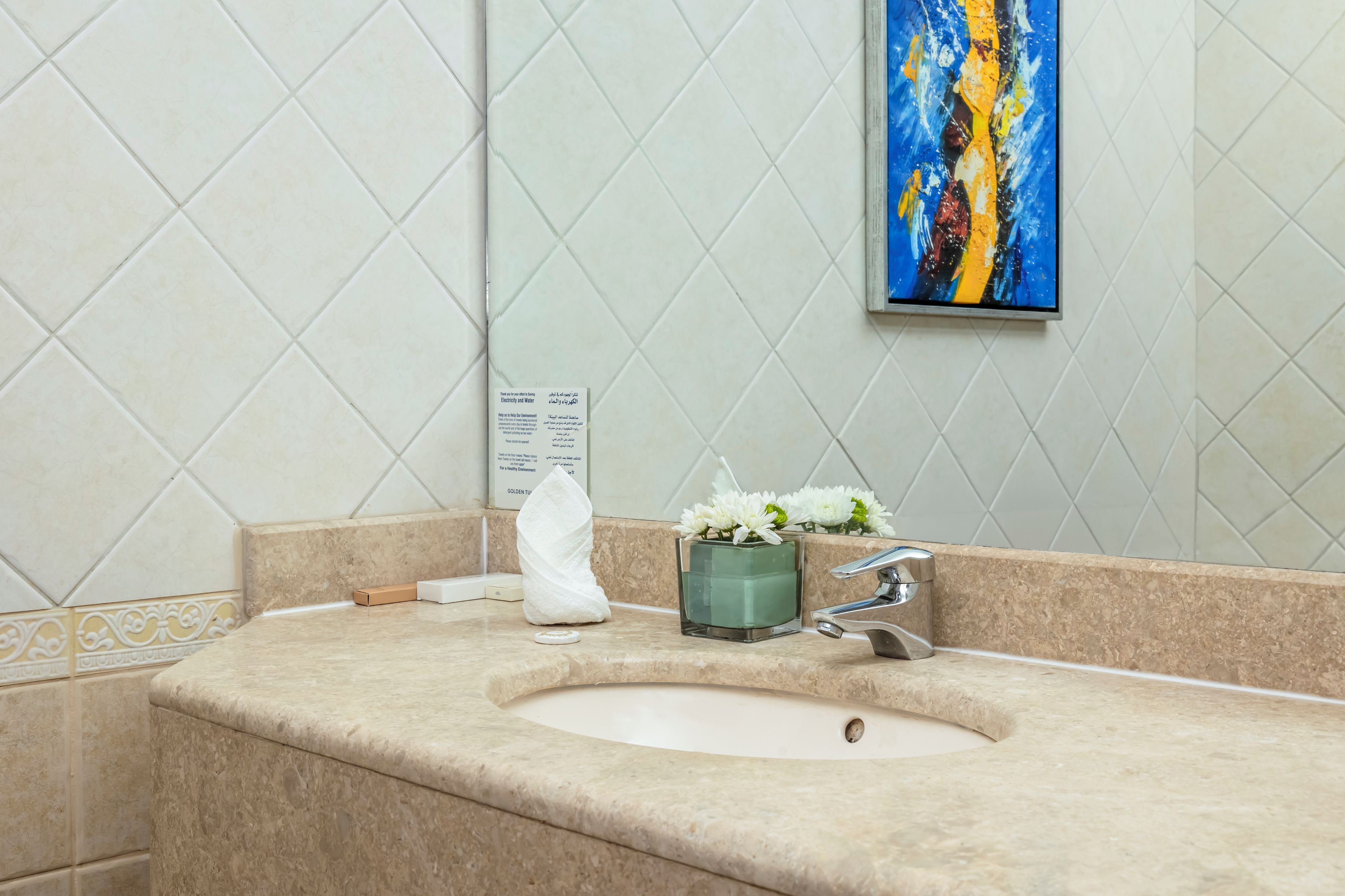 A bathroom with a sink, a towel, a glass vase with flowers, and a painting on the wall.