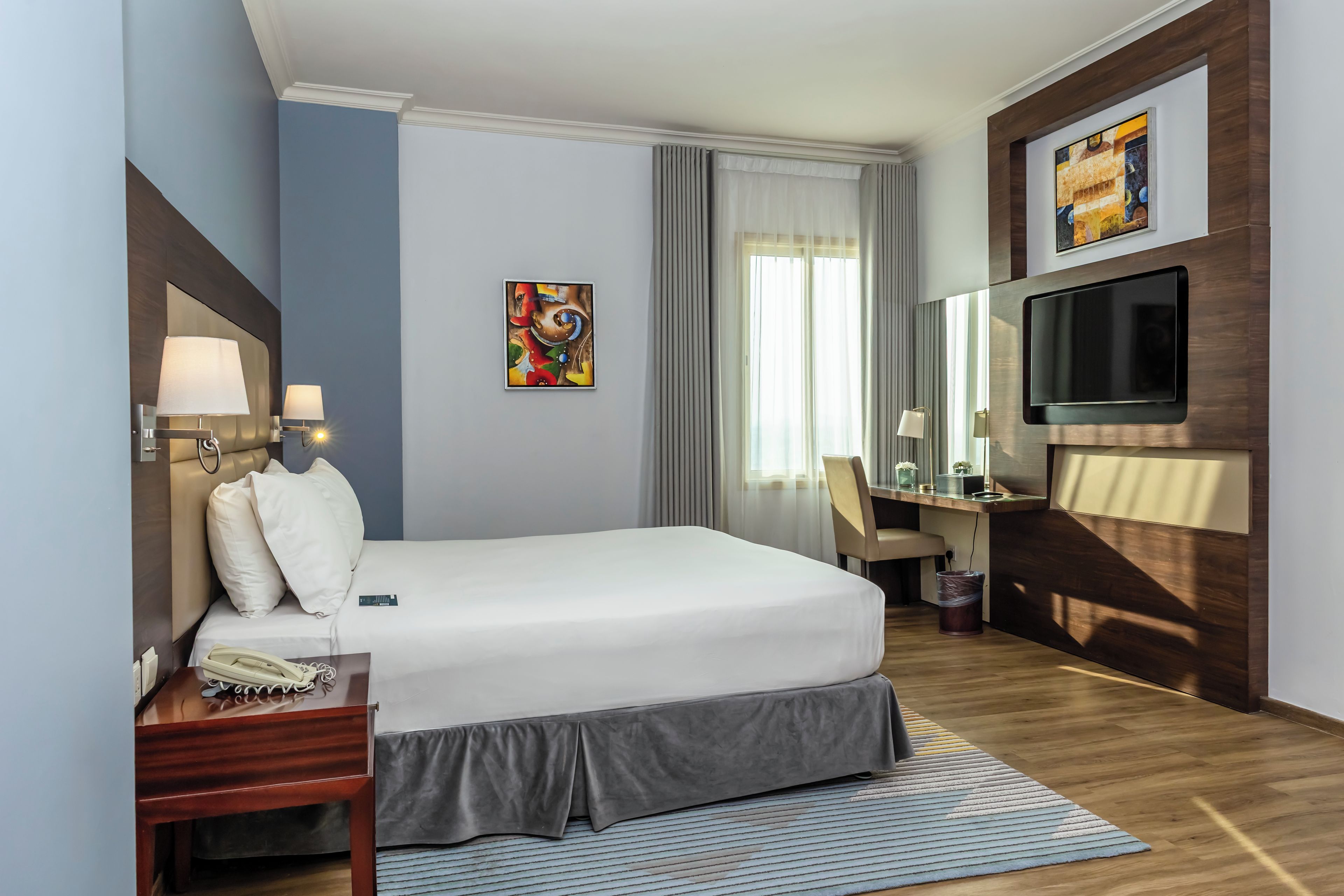A hotel room with a bed, bedside table, lamp, desk, chair, TV, and artwork.