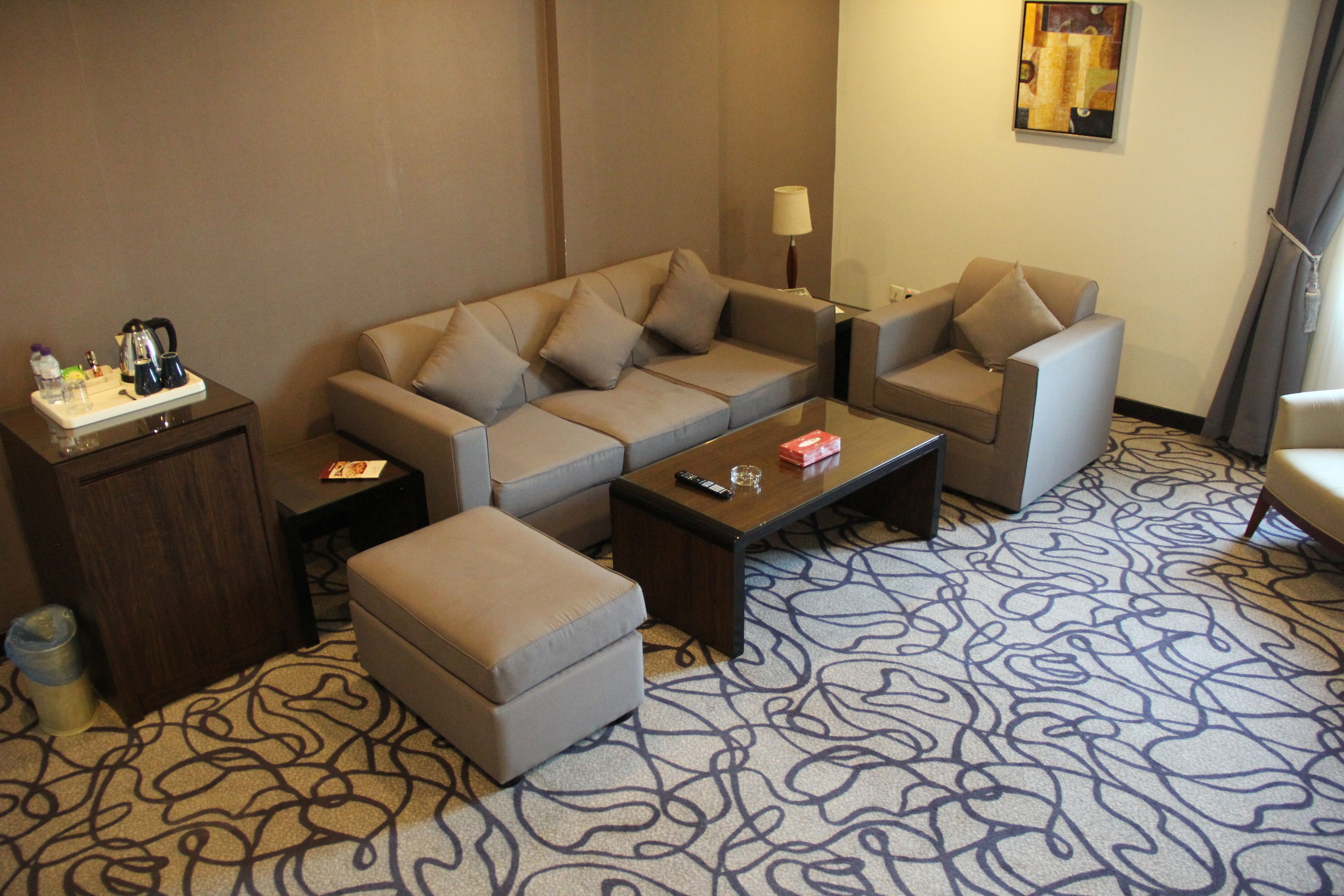 A hotel room with a sofa set, a coffee table, a side table with a lamp, and a patterned carpet.