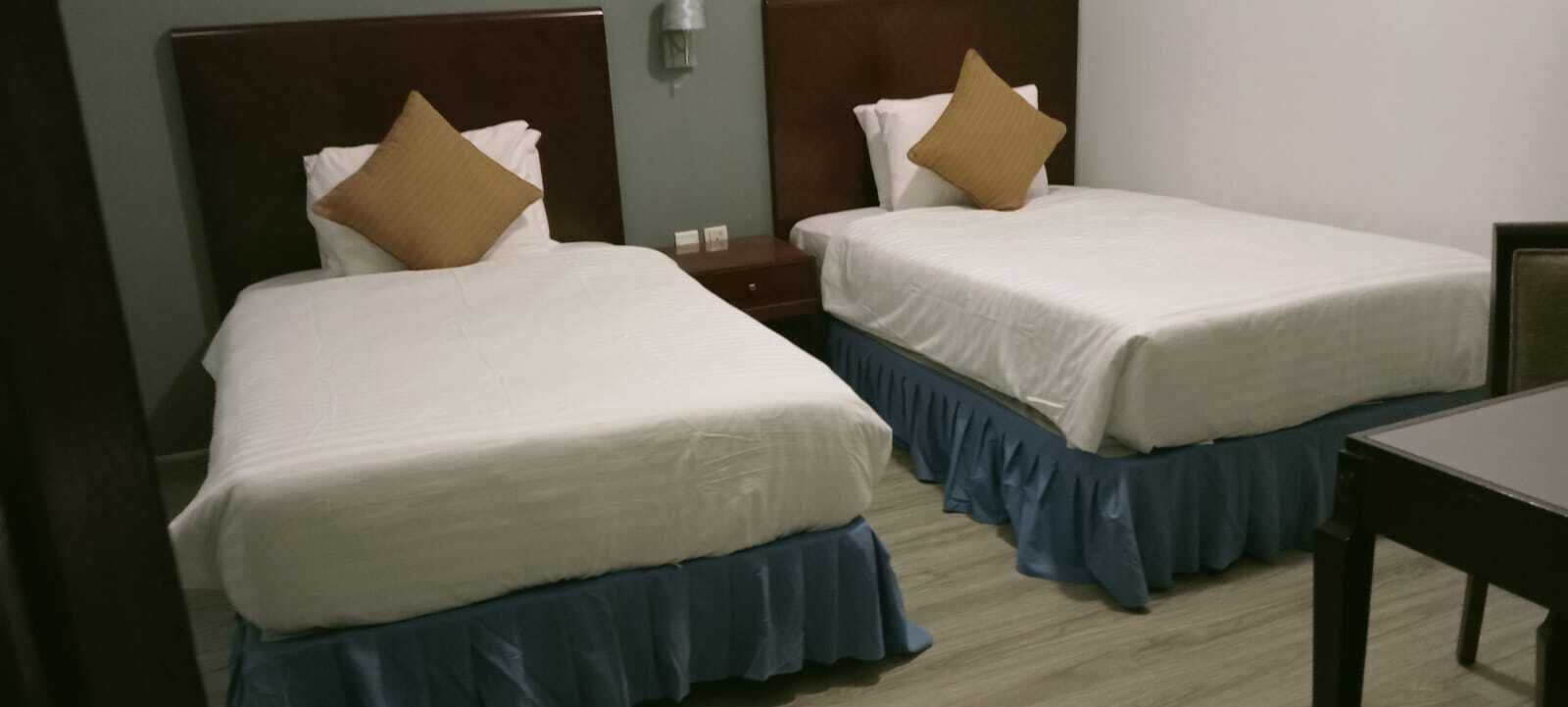 Deux lits simples avec des têtes de lit, une table de chevet, une table et une lampe fixée au mur.