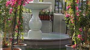 A fountain with a white pedestal, surrounded by pink flowering plants and potted trees, set against a brick pavement and a metal fence. - Golden Tulip Al Khobar (Al Khobar)